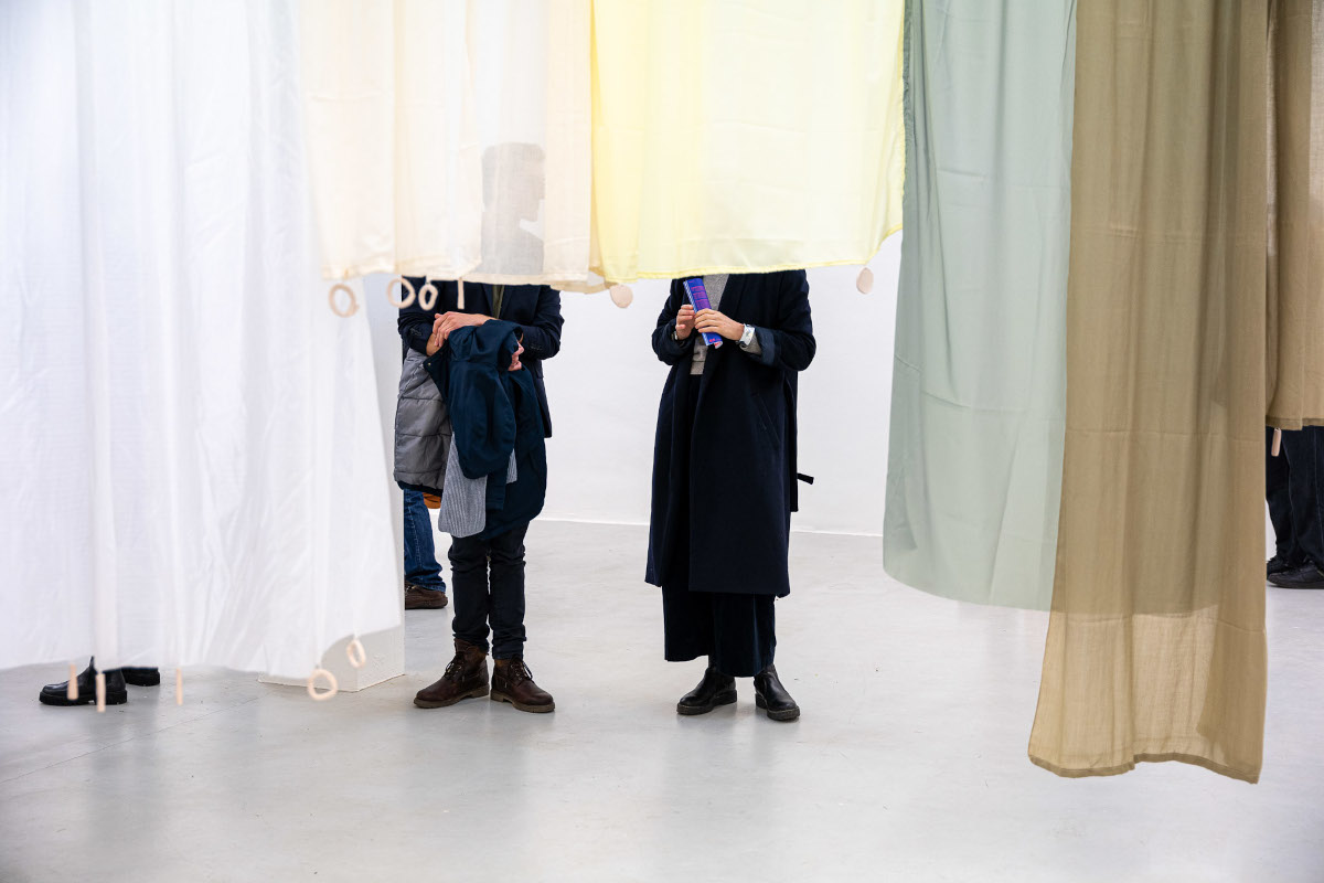 Ausstellungsansicht, Menschen im Gespräch, halb verdeckt von der Stoff/Vorhanginstallation von Martina Boettiger