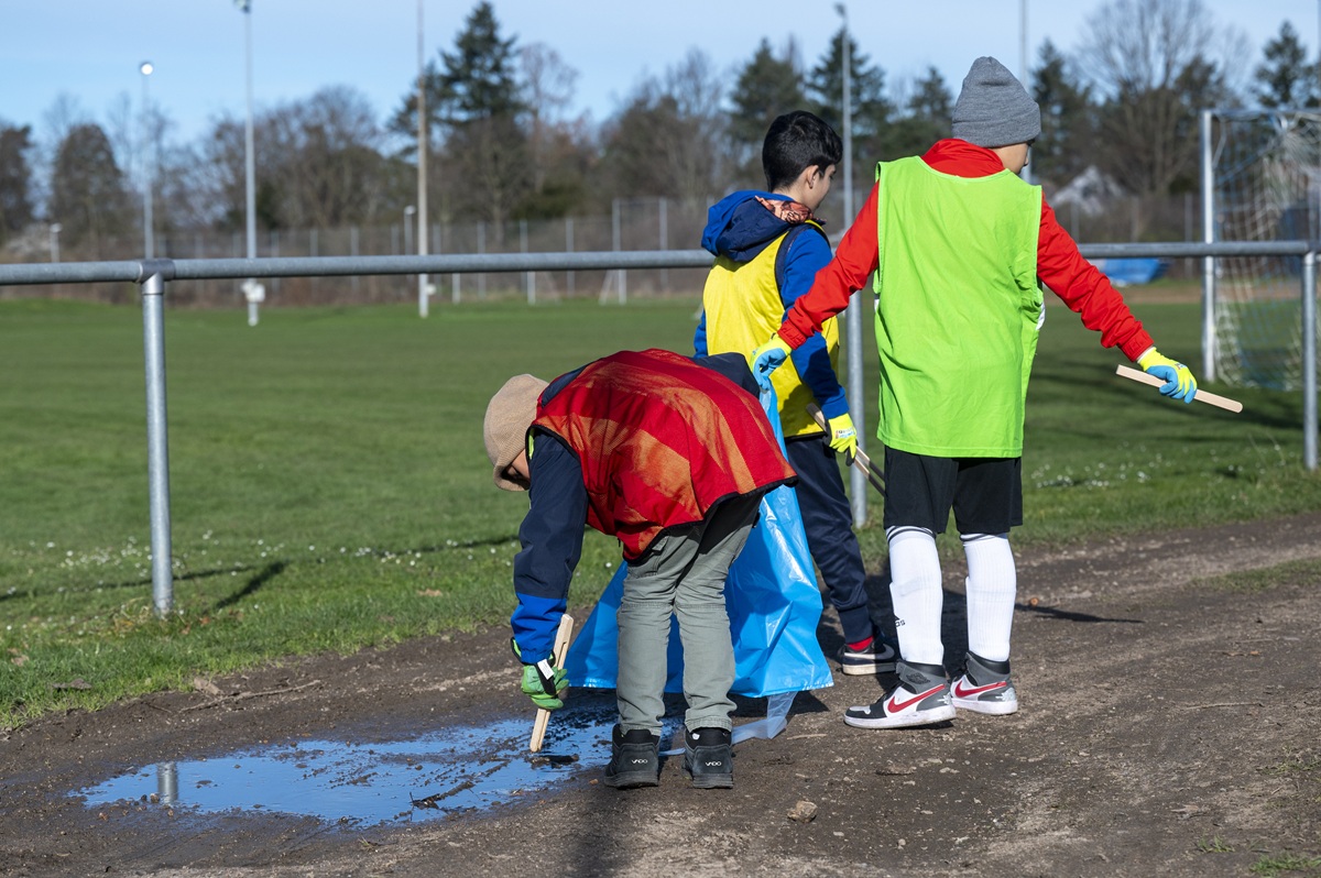 Drei Schüler stehen neben einem Fußballplatz. Der ganz links bückt sich, die anderen zwei deuten mit Holzzangen auf den Boden. Sie stehen mit dem Rücken zur Kamera.