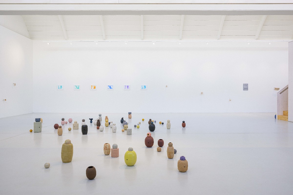 Ausstellungsansicht mit zahlreichen kleinen keramischen Skulpturen in organischen Formen und gedeckten Farben, frei auf dem Boden eines hellen Galerieraums verteilt von Tanja Götzmann, an der Wand kleinformatige grafische Arbeiten von Tim Bohlender und kleine holzreliefs von Nozomi Tanabe