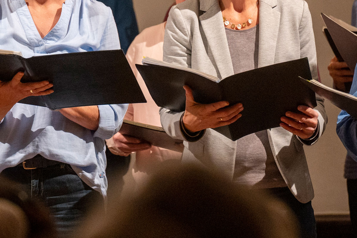 mehrere Personen halten Noten in der Hand vor einem Publikum, Ausschnitt ohne Gesichter