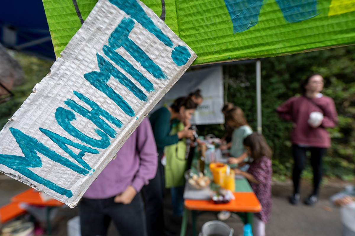 Ein Schild mit blauer Schrift "Mach Mit!", im Hintergrund verschwommen ein Waffelverkauf