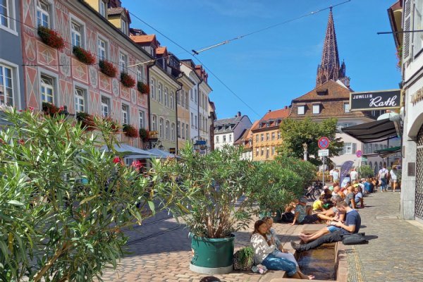 Historische Häuser mit Münster und gepflasterte Straße und Pflanzkübeln, Menschen sitzen mit den Füßen im Bächle