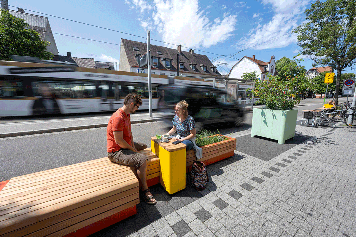 Zwei Menschen sitzen auf einer Bank auf einem ehemaligen Parkplatz.