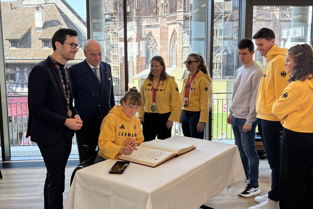 Die paralympischen Gewinner*innen sind mit OB Horn und Gundolf Fleischer im Café Inklusiv um einen Tisch versammelt, an dem sich die Paralympische Sportlerin Anna-Lena Forster gerade in das Goldene Buch der Stadt einträgt.