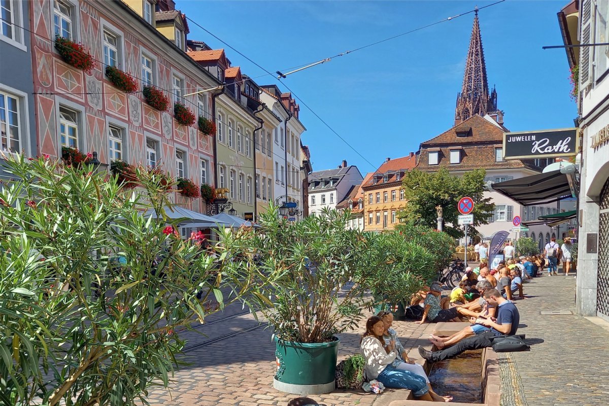 Historische Häuser mit Münster und gepflasterte Straße und Pflanzkübeln, Menschen sitzen mit den Füßen im 