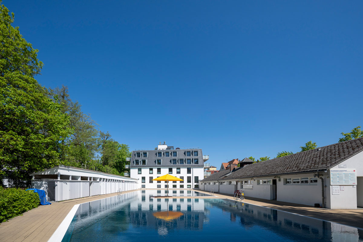 Mittig ein Schwimmbecken, im Hintergrund ein Gebäude, Rechts und Links Umkleidekabinen