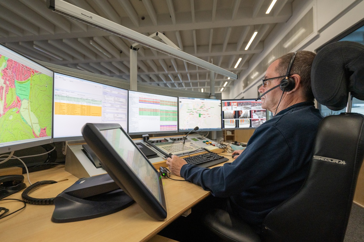 Ein Mann von der Leitstelle sitzt an einem Schreibtisch mit vielen Computerbildschirmen und nimmt mit einem Headset Anrufe entgegen.