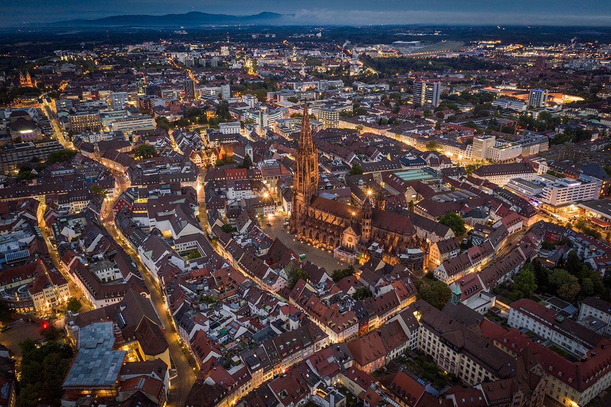 Luftaufnahme von Freiburg im Breisgau. Freiburger Münster in der Mitte des Rahmens. Beleuchtete Straßen und Gebäude im Morgengrauen. Baden-Württemberg in Deutschland