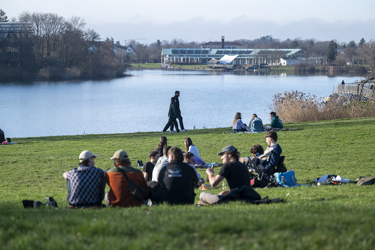 Eine Gruppe Menschen sitzt bei Frühlingswetter am See im Seepark.