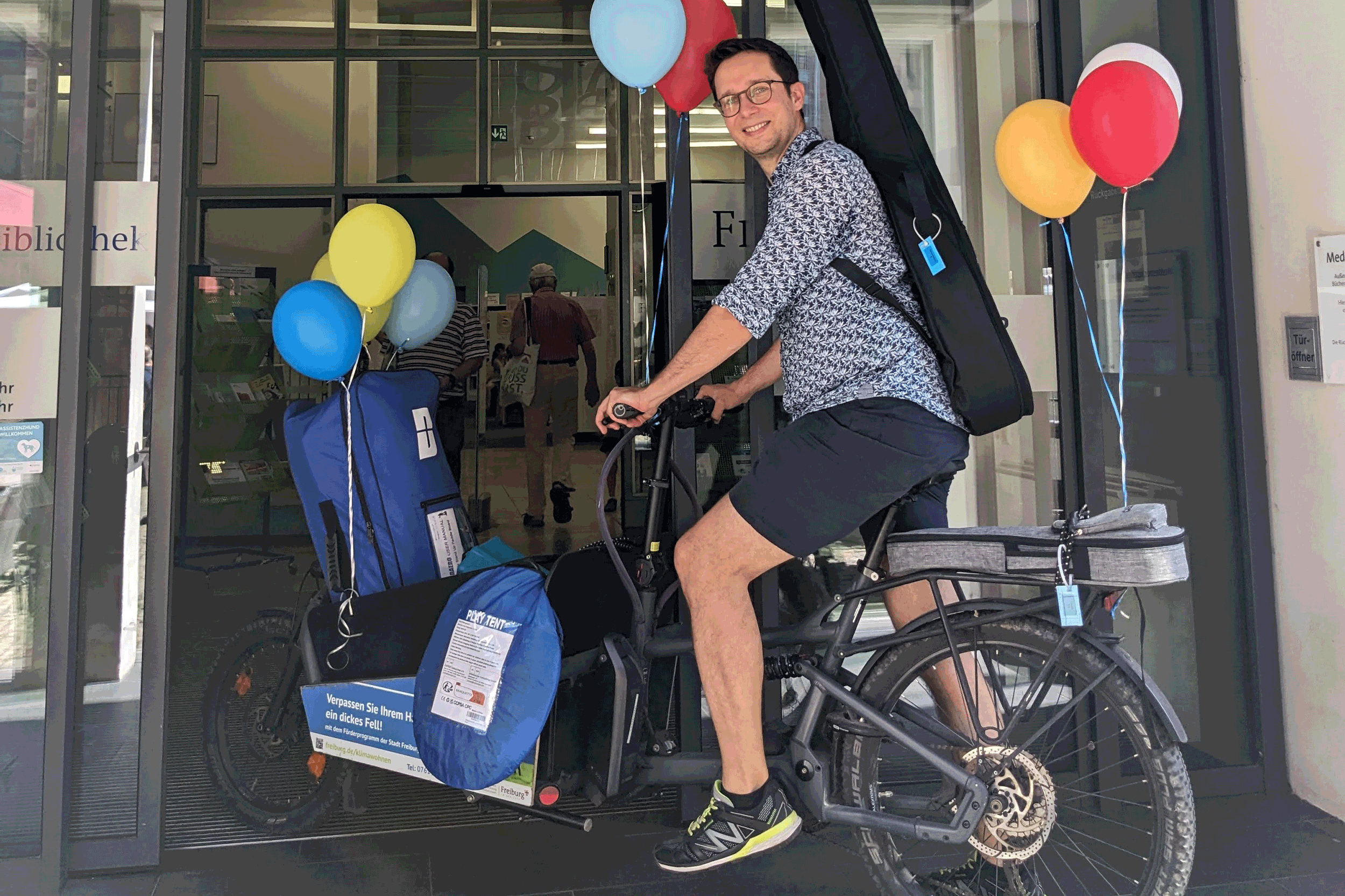 Mensch auf Rad vollgepacktem Lastenrad mit Luftballons 