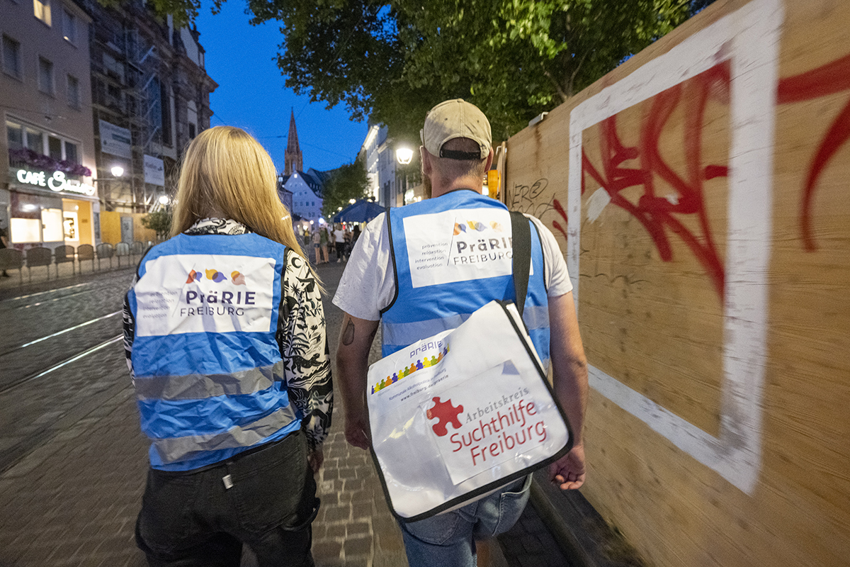 Zwei junge Menschen laufen nachts durch Freiburg. Sie tragen blaue Warnwesten mit dem Logo von Prärie. Eine Person trägt eine Tasche von der Suchthilfe Freiburg.