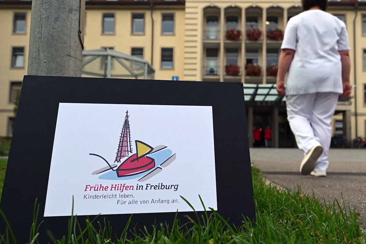 Pflegepersonal läuft vor Klinikeingang, Schild mit Aufschrift: Frühe Hilfen in Freiburg