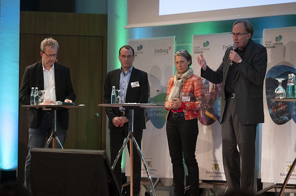 Drei Männer und eine Frau mit Mikrofonen stehen auf Podium