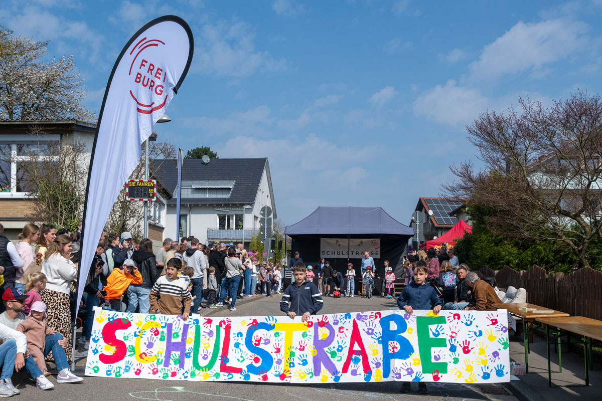 Straße auf der viele Kinder und Erwachsene sind, ein buntes Banner mit der Aufschrift Schulstraße